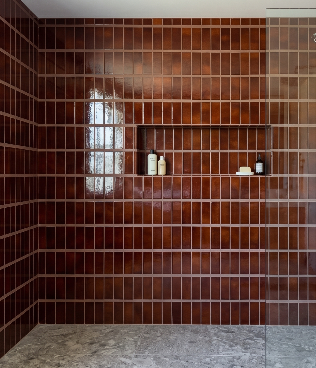 A modern shower wall featuring glossy, dark reddish-brown rectangular tiles arranged in a grid pattern, with a recessed shelf holding a few small toiletry bottles. The floor is a light gray stone tile, and a glass shower door is visible on the right side.