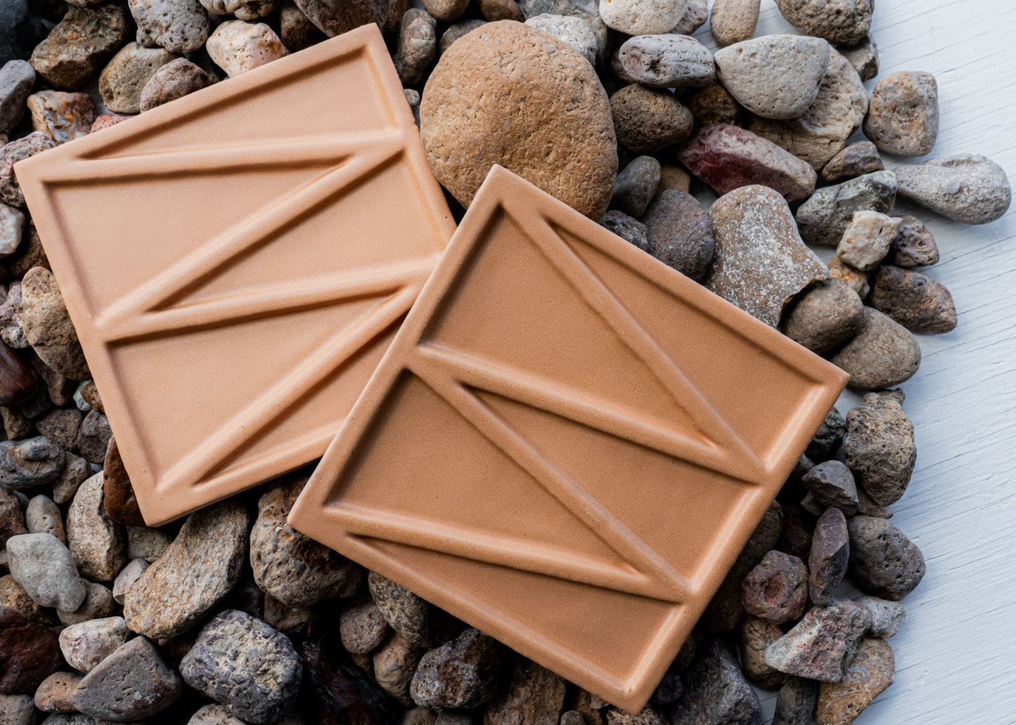 Two terracotta solid tiles by Clay Imports, featuring a raised geometric pattern and placed on pebbles.