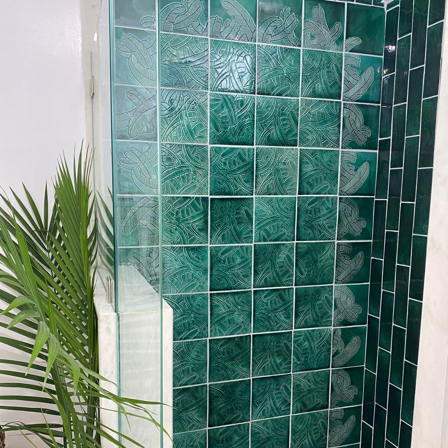 Dark green Platanos Forest 6x6 tiles by Clay Imports, featuring embossed tropical leaf patterns on a shower wall with a palm plant.