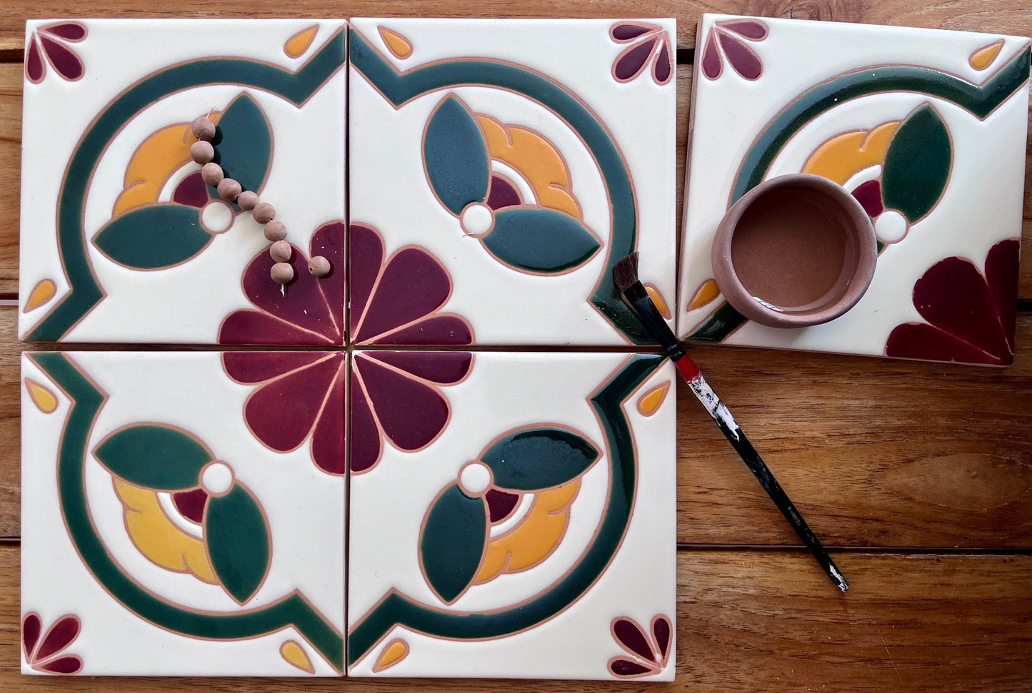 White relief Talavera tile by Clay Imports, featuring an autumnal green, yellow, and maroon floral design, arranged with other tiles, beads, and painting supplies.