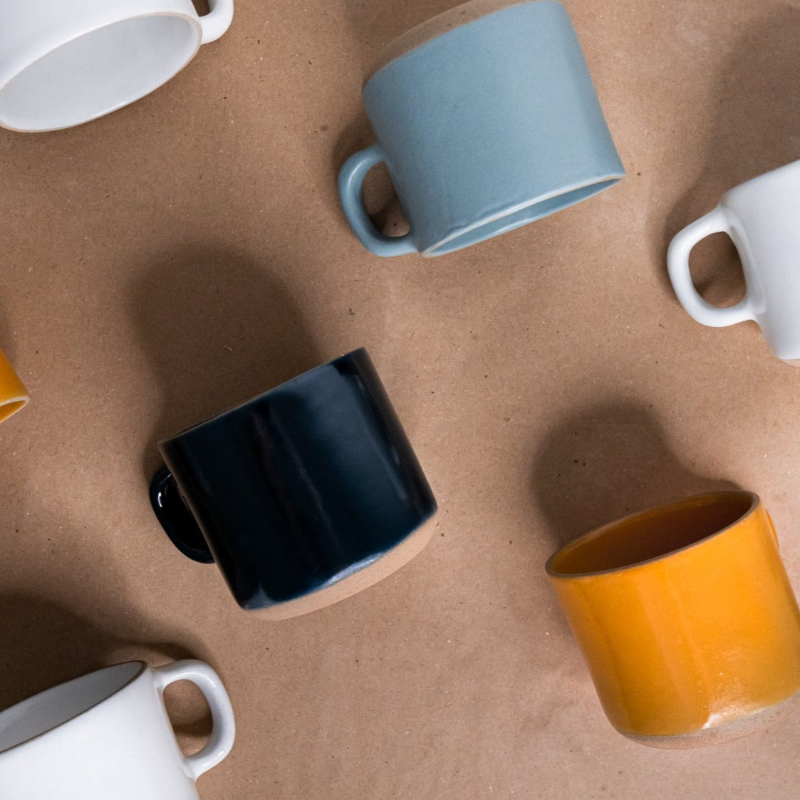 Assortment of smooth ceramic Essential Mugs by Clay Imports in various colors scattered on brown paper.