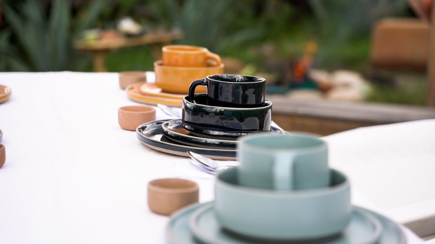 Stacked ceramic Everything Bowls in dark green and teal by Clay Imports, on an outdoor table setting with other bowls and plates.