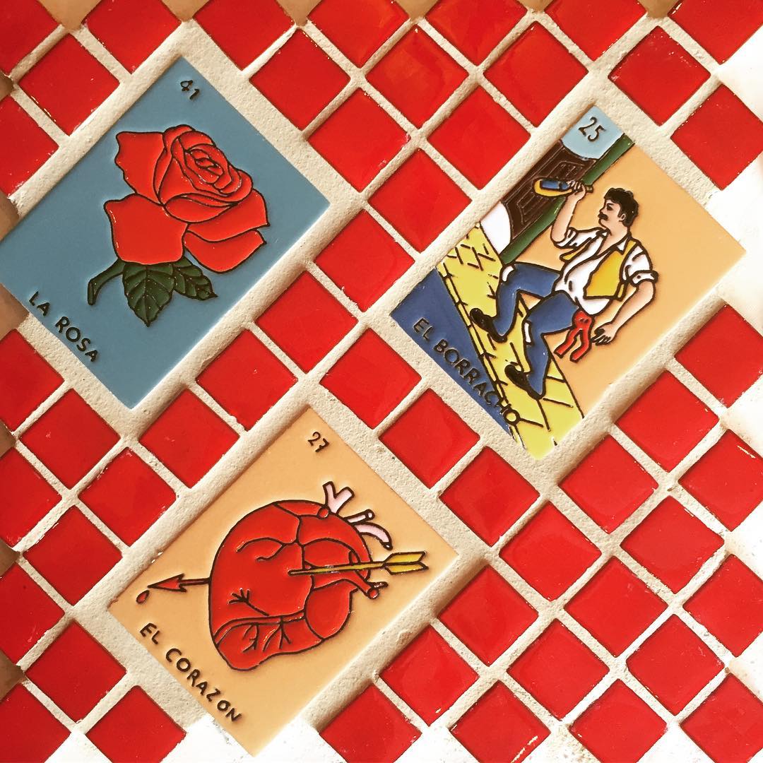 Original Lotería Set by Clay Imports, featuring three ceramic game tiles ('La Rosa', 'El Borracho', 'El Corazón') set in a red mosaic.