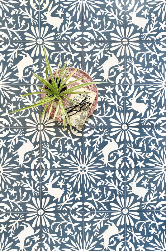 Charming blue and white patterned tiles with rabbits and flowers, styled with a potted plant, book, and reading glasses.