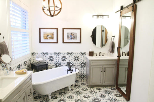 A bright, stylish bathroom featuring ornate patterned tiles, a classic clawfoot tub, barn door mirror, and light gray vanities.