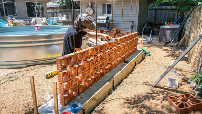 A person carefully levels a new row of orange breeze blocks on a concrete foundation in a backyard for a DIY wall.