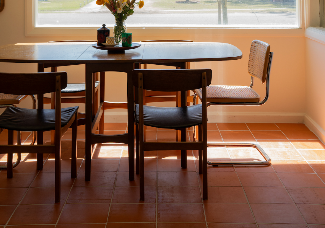 Sun-drenched dining area featuring a wooden table, mixed chairs, and warm terracotta floor tiles.