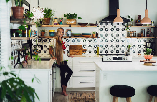 A smiling woman stands proudly in her stylish, intentionally designed kitchen featuring a striking black and white sustainable tile backsplash.