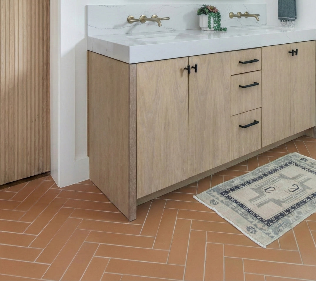 Sophisticated bathroom with a light wood double vanity, white marble top, elegant brass fixtures, and warm terracotta herringbone floor.