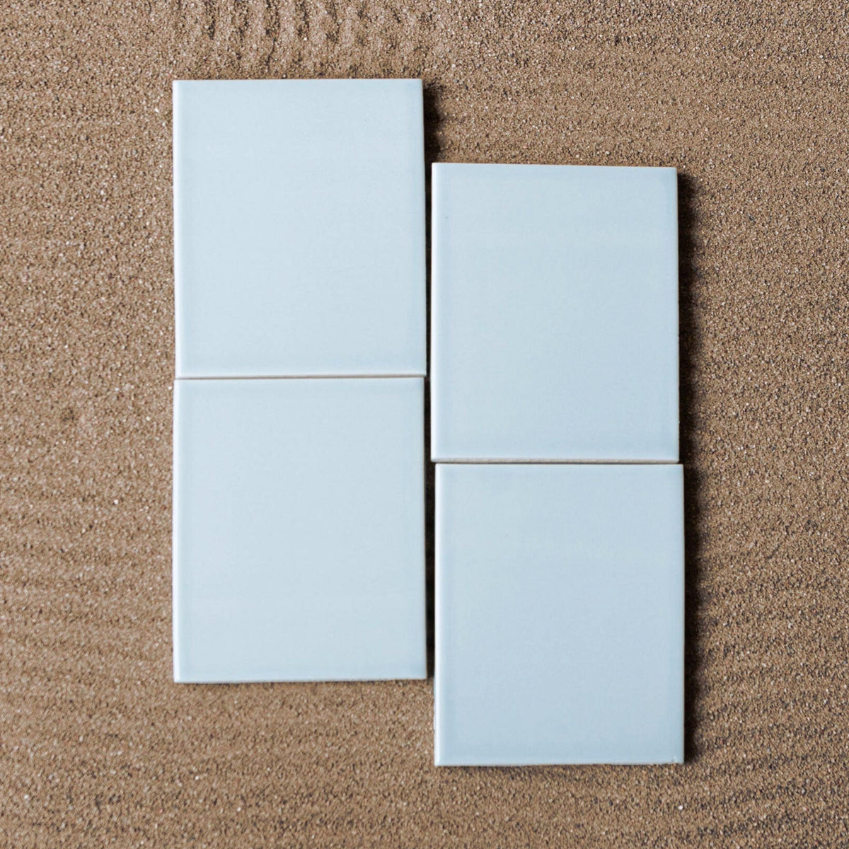 Light blue glossy 4x5 solid tiles by Clay Imports, arranged in a grid on textured sand with raked patterns.