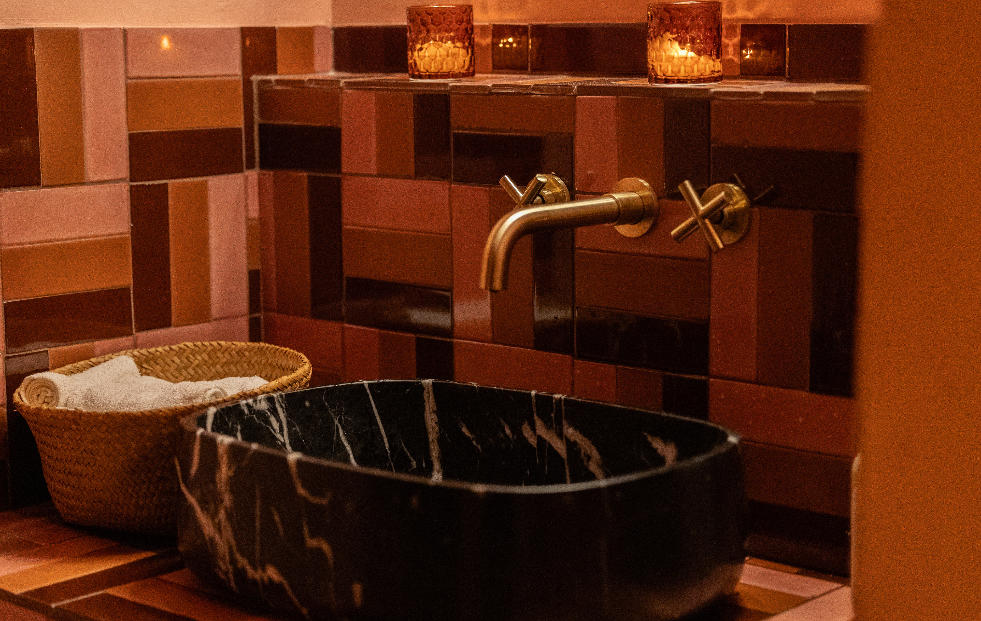 Warm, intimate bathroom featuring black marble vessel sink, brass faucet, and terracotta-toned geometric tiles, illuminated by soft candle glow.