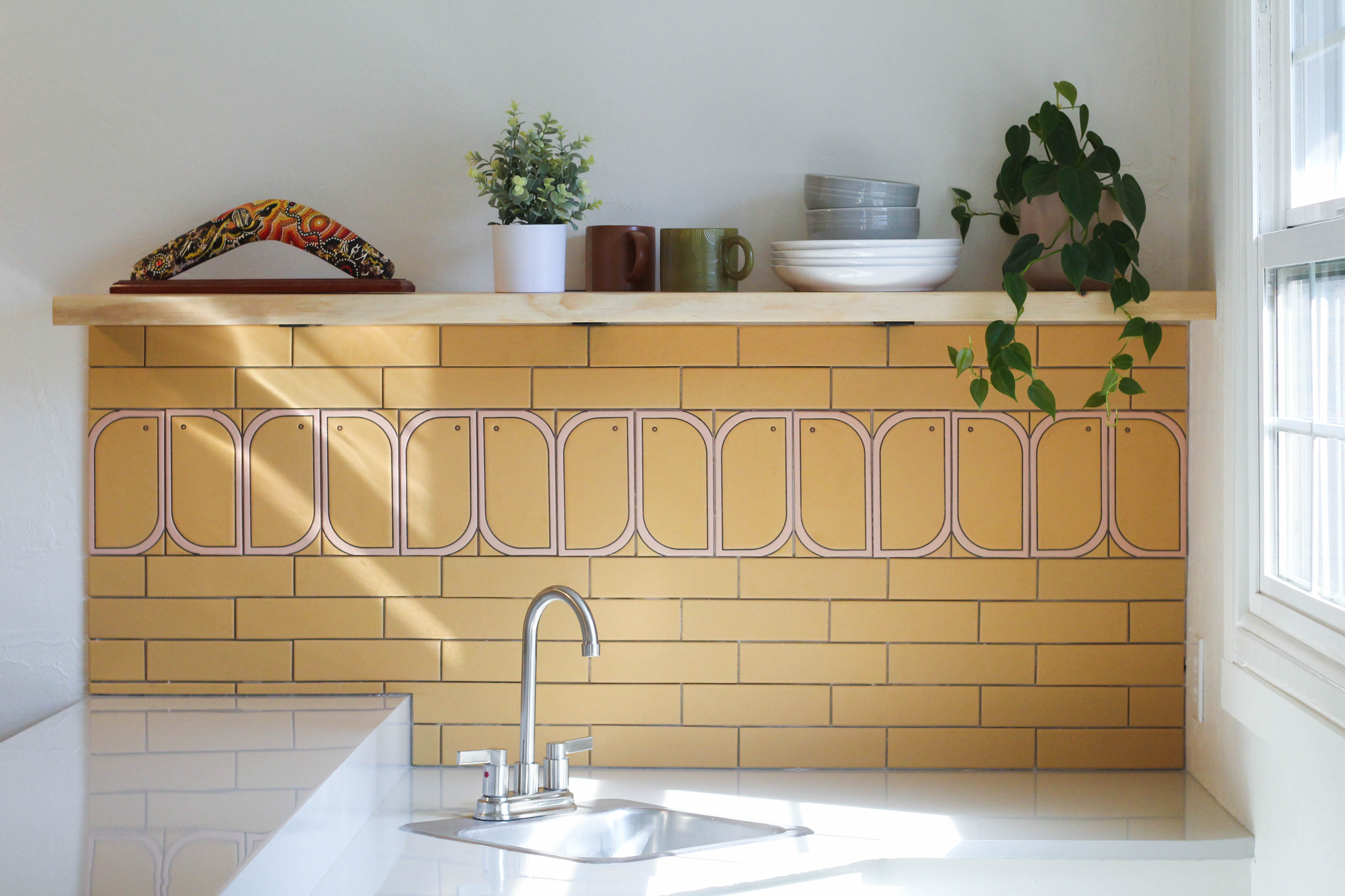 Warm, sunlit kitchen featuring a distinctive yellow and pink arched tile backsplash, showcasing modern dishware and greenery.