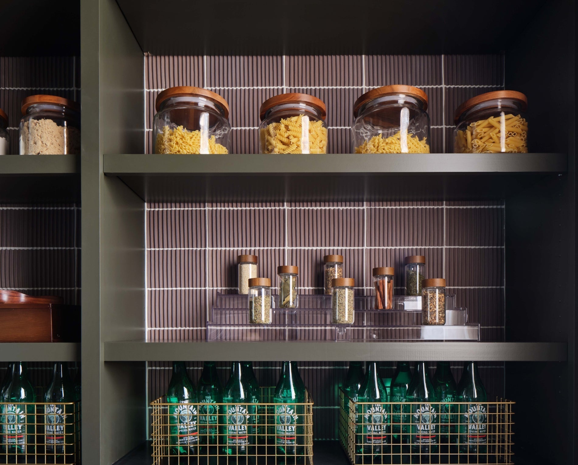 Organized pantry with warm wood-lidded jars, spices, and green bottles on olive shelves against earthy ribbed tiles, embodying smart storage.