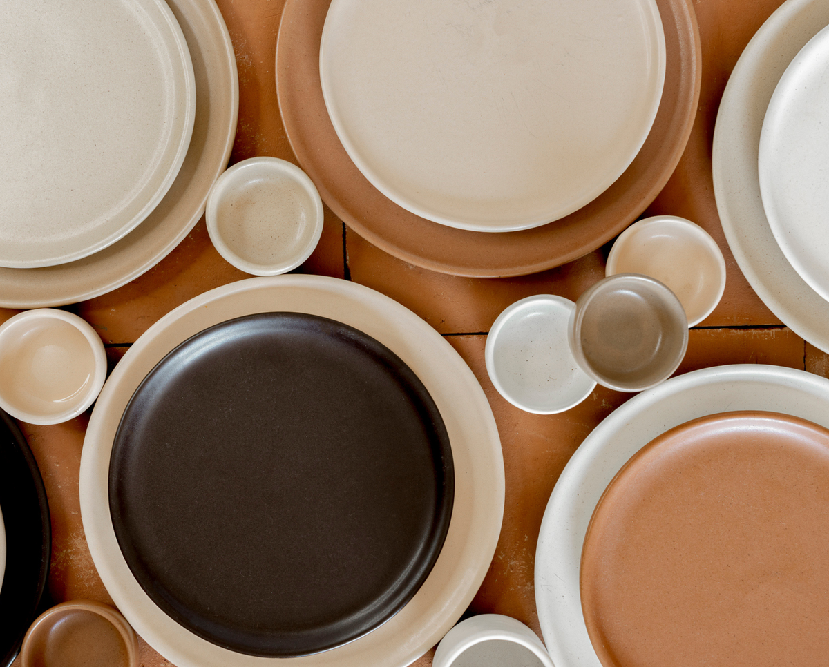 Modern Poco x Clay ceramic dinnerware stacks in vibrant orange, deep black, warm mustard, and cool teal against an earthy background.