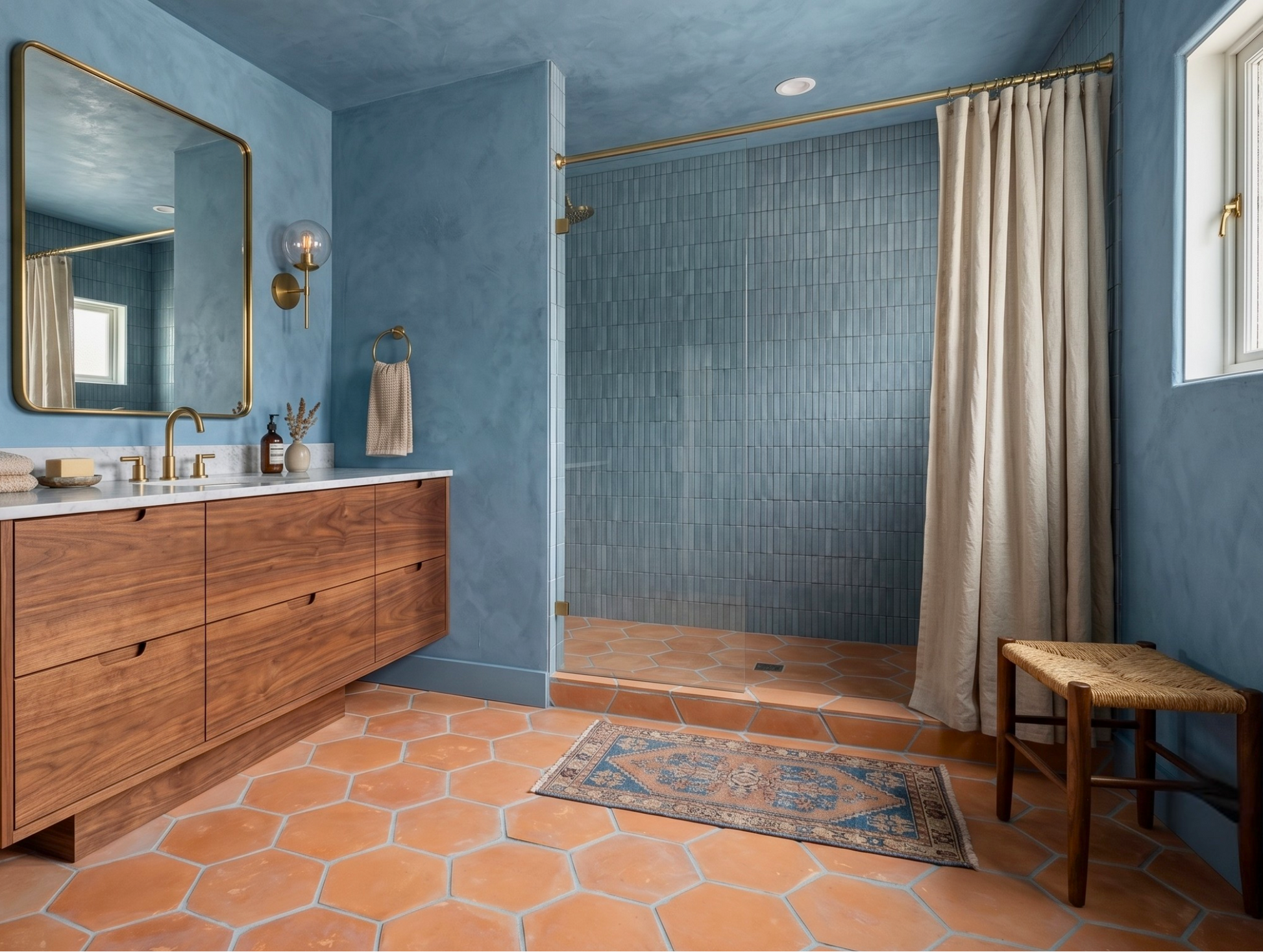A modern bathroom interior with blue walls, a wood vanity with a white countertop and mirror, terracotta hexagonal tile flooring, a tiled shower area with a beige curtain, and a small wooden stool.