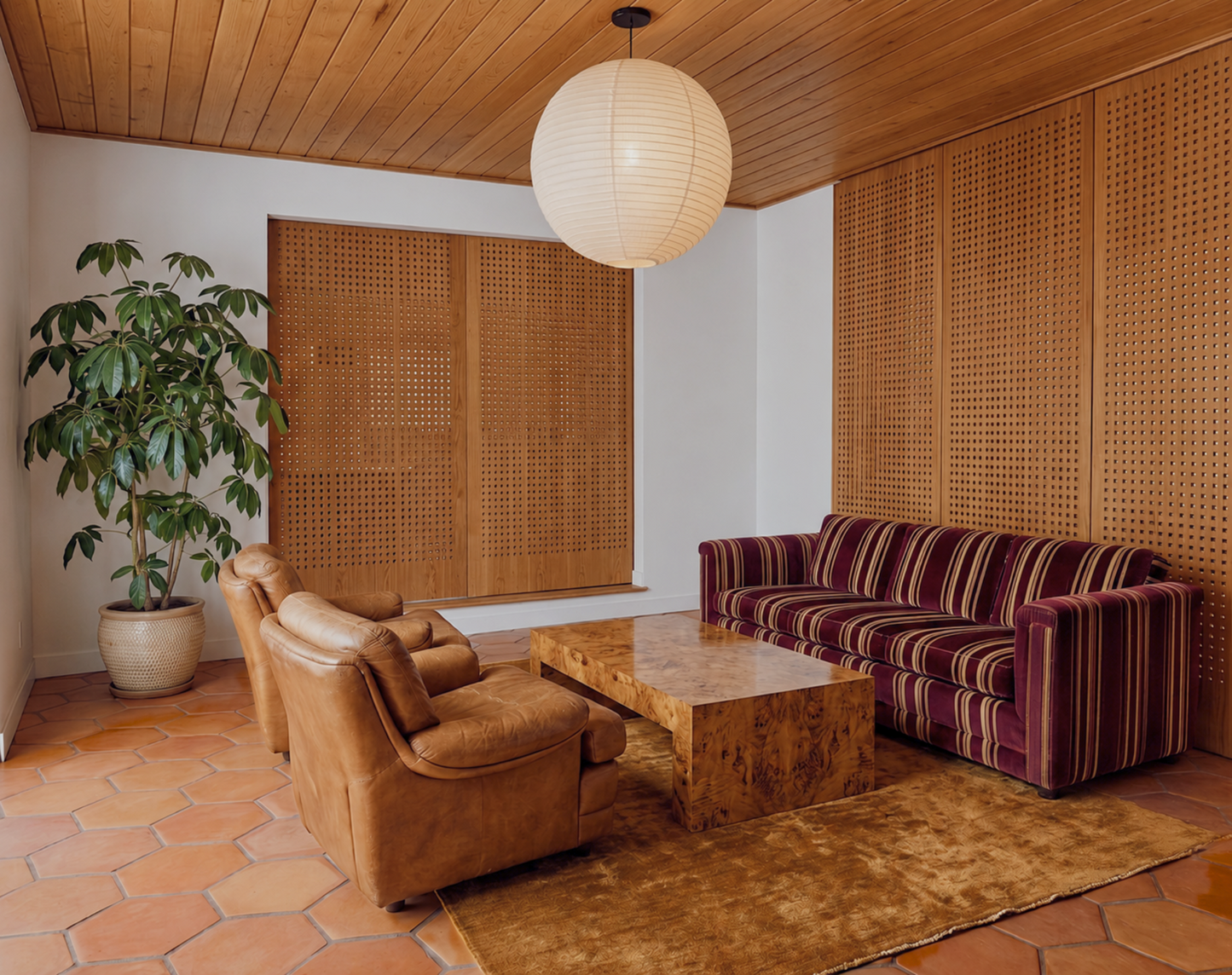 A retro-style living room featuring a brown leather armchair, a purple and brown striped velvet sofa, a wooden coffee table, terracotta hexagonal floor tiles, and a large potted plant.