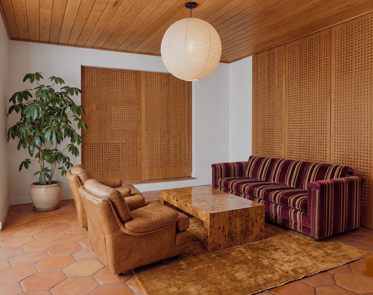 A retro-style living room featuring a brown leather armchair, a purple and brown striped velvet sofa, a wooden coffee table, terracotta hexagonal floor tiles, and a large potted plant.
