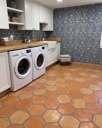 A bright laundry room with terracotta hexagonal floor tiles, white cabinets, a double-front washer and dryer, a farmhouse sink, and blue floral wallpaper.