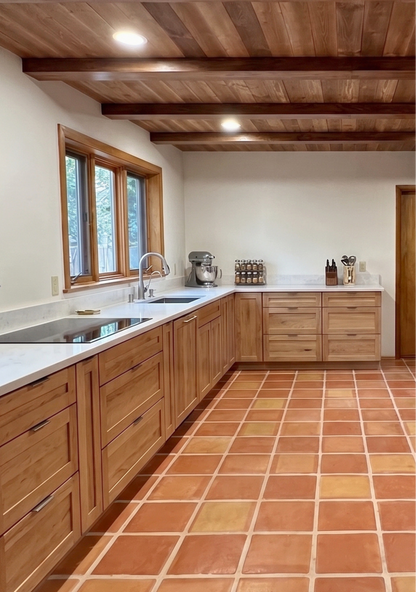 A modern kitchen interior with light oak wood cabinets, white marble countertops, a double sink, a wood-framed window, and terracotta tile flooring.