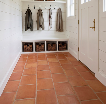 A bright, clean entryway with terracotta tile flooring, white shiplap walls, a white bench with four woven storage baskets, hanging coats and scarf, and a white door with a brass handle.