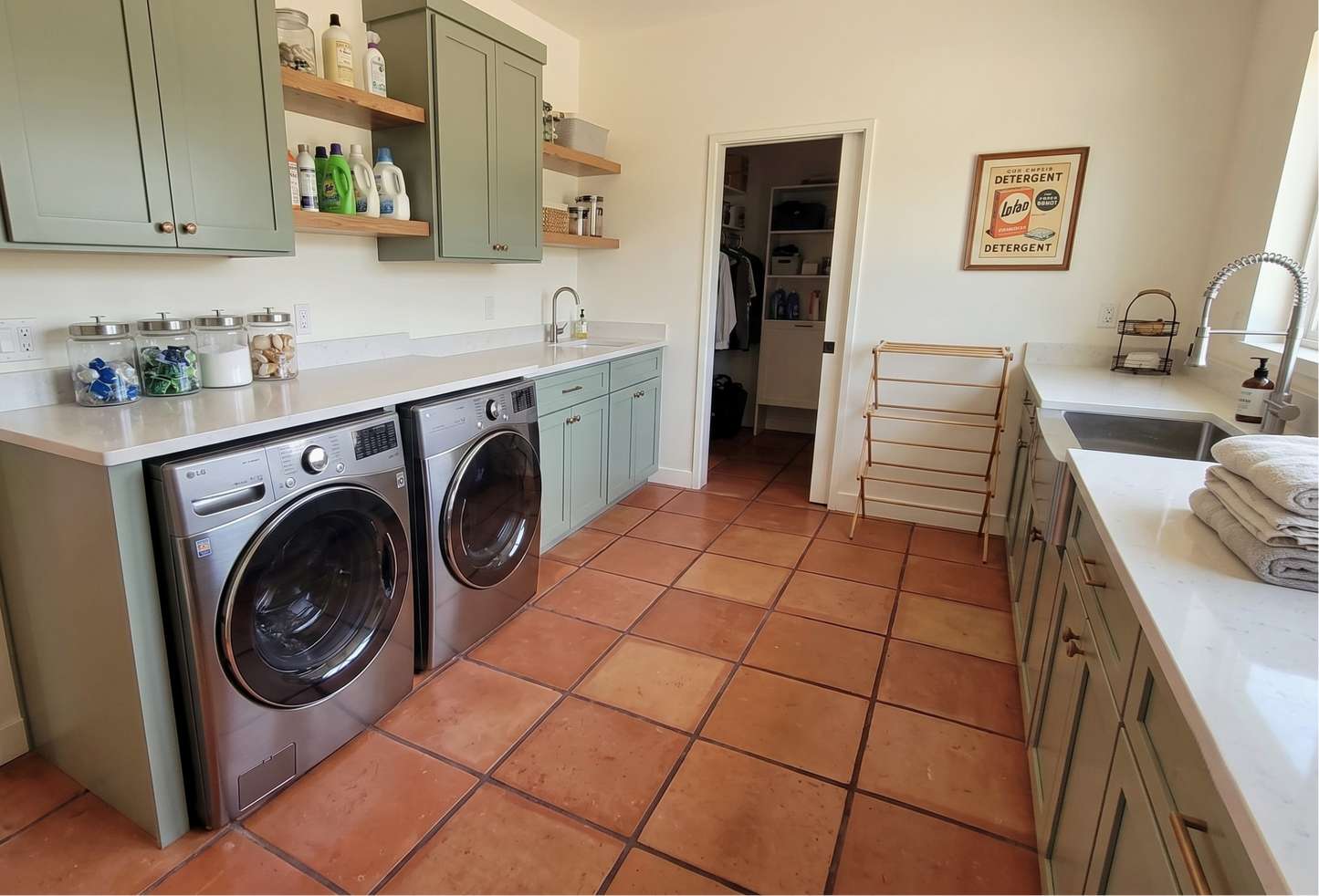A bright utility room with two stainless steel front-load washing machines under sage green cabinets, terracotta tile floors, white countertops, and a sink area with folded towels.