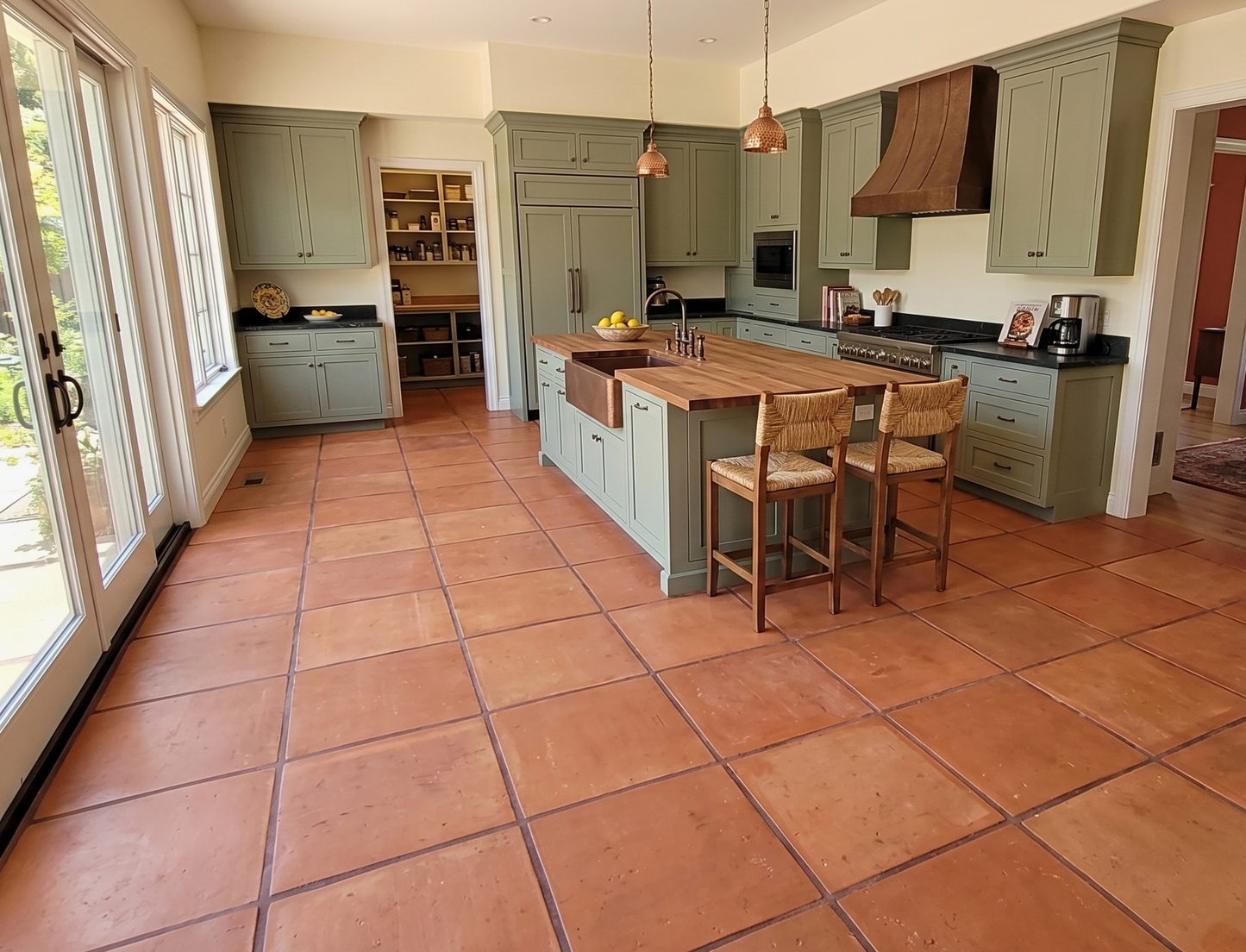 A spacious kitchen with sage green cabinets, terracotta tile flooring, and a central island with a wood countertop and two wooden bar stools.