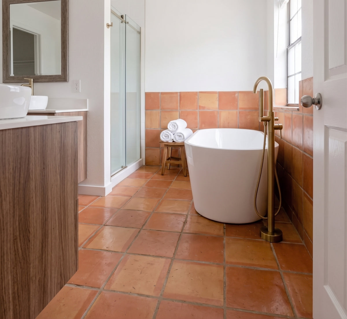 A modern freestanding bathtub with a tall brass faucet and handheld shower attachment in a bright bathroom with terracotta tile floors and walls, a glass shower door, and a white vanity.