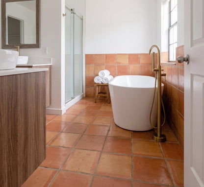 A modern freestanding bathtub with a tall brass faucet and handheld shower attachment in a bright bathroom with terracotta tile floors and walls, a glass shower door, and a white vanity.
