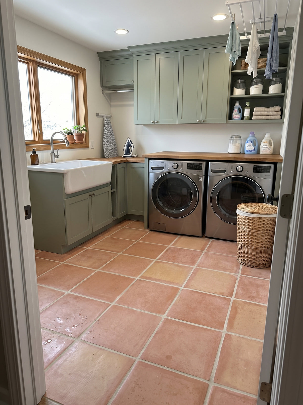 A spacious modern laundry room features sage green cabinets, stainless steel washer and dryer, and terracotta tile flooring.