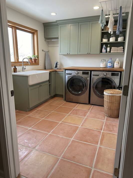 A spacious modern laundry room features sage green cabinets, stainless steel washer and dryer, and terracotta tile flooring.