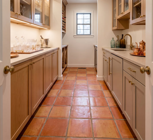 A long narrow kitchen or pantry corridor with light wood cabinets, cream countertops, brass hardware, and terracotta tile flooring. A small window is centered in the back wall, and bar glasses, bottles, and copper mugs are arranged on the counters.