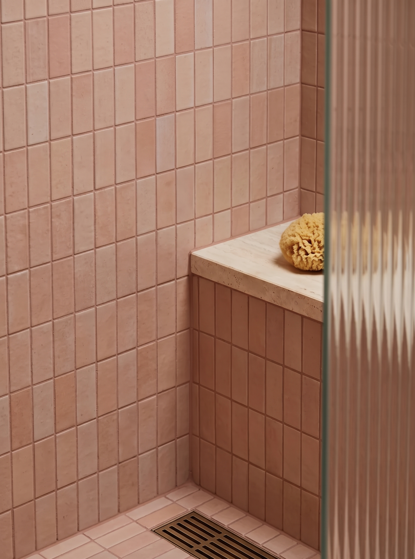 A close-up of a tiled shower corner with warm peach-pink square tiles, a light stone bench, a natural loofah sponge, and a glass door with vertical ribbed texture on the right.