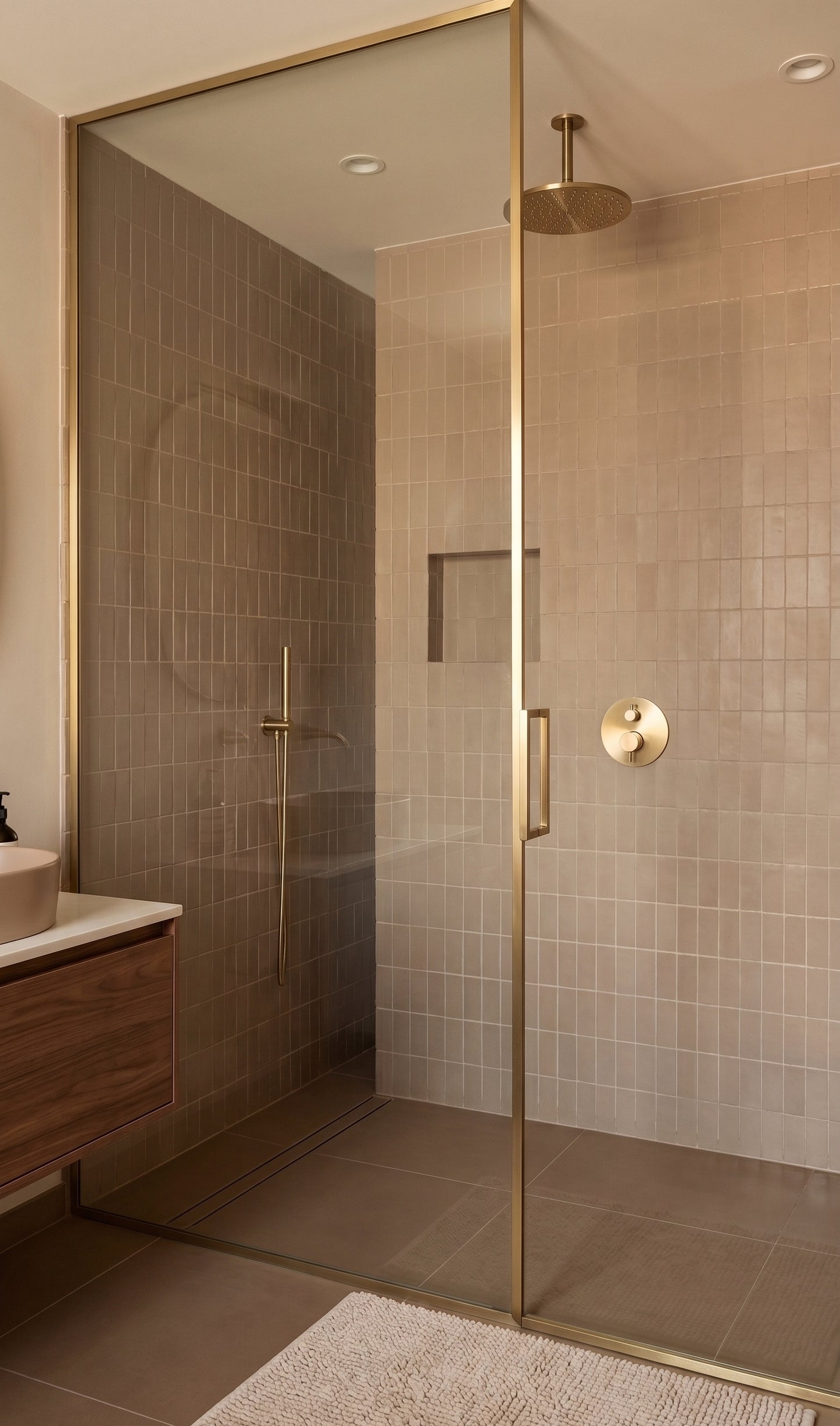 A modern walk-in shower enclosure with a frameless glass door and brushed brass hardware, set in a bathroom with beige tiled walls and floor.