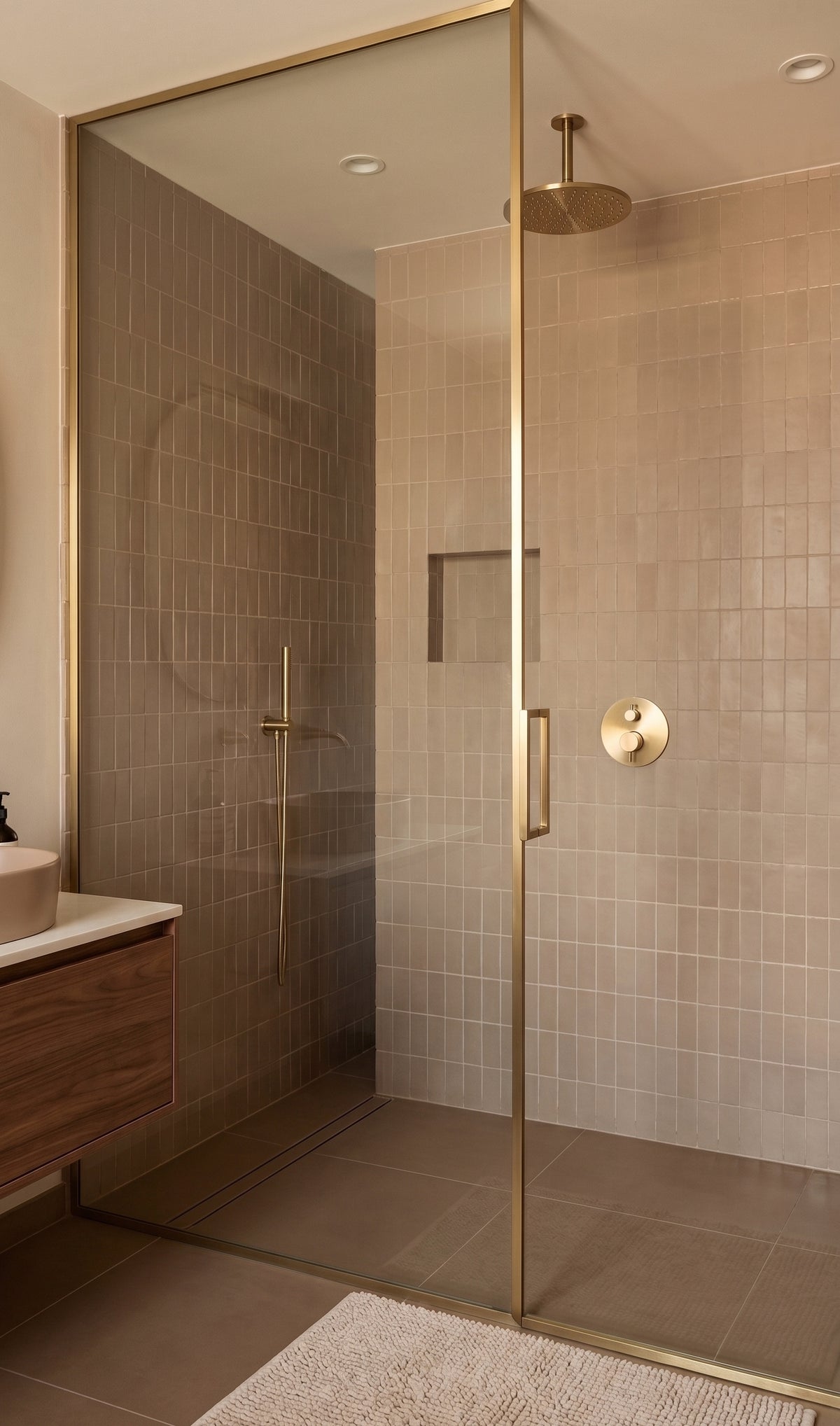 A modern walk-in shower enclosure with a frameless glass door and brushed brass hardware, set in a bathroom with beige tiled walls and floor.