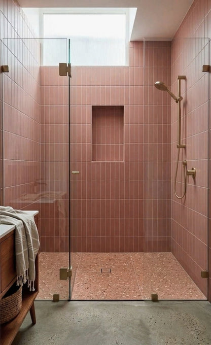 A modern walk-in shower features pink vertical tiled walls, a terrazzo floor, and elegant brass fixtures under a skylight.