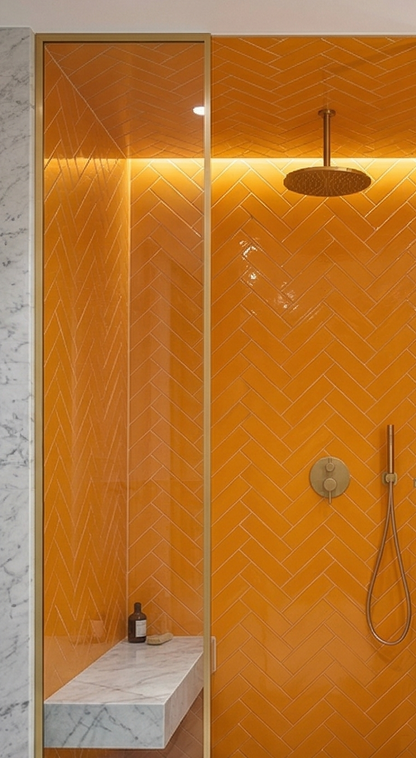 Marigold solid tile by Clay Imports in a glossy herringbone pattern, installed in a modern shower with gold fixtures and a marble bench.