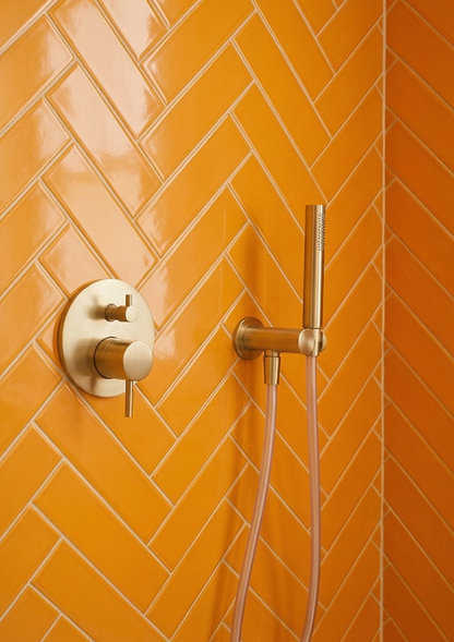Marigold 2x6 Solid Tile by Clay Imports, in a glossy orange herringbone pattern with brushed gold shower fixtures.