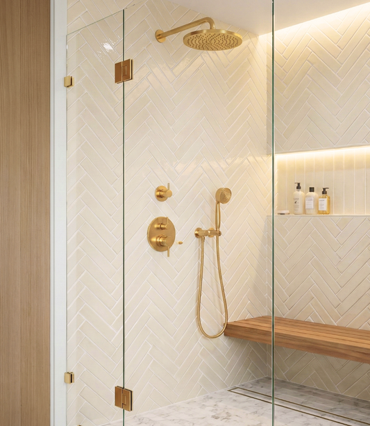 Fossil Gloss 2x12 Solid Tile by Clay Imports, light beige glossy herringbone pattern on a shower wall with gold fixtures and wooden bench.