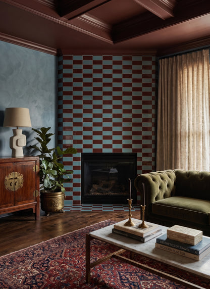 A cozy living room features a striking fireplace surround tiled in red and blue brick patterns, complemented by an olive green tufted sofa and a reddish-brown coffered ceiling.