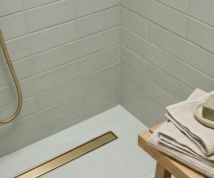 Light green matte rectangular solid tile by Espadin, installed in shower corner with white grout, gold linear drain, and beige waffle towels on wooden stool.