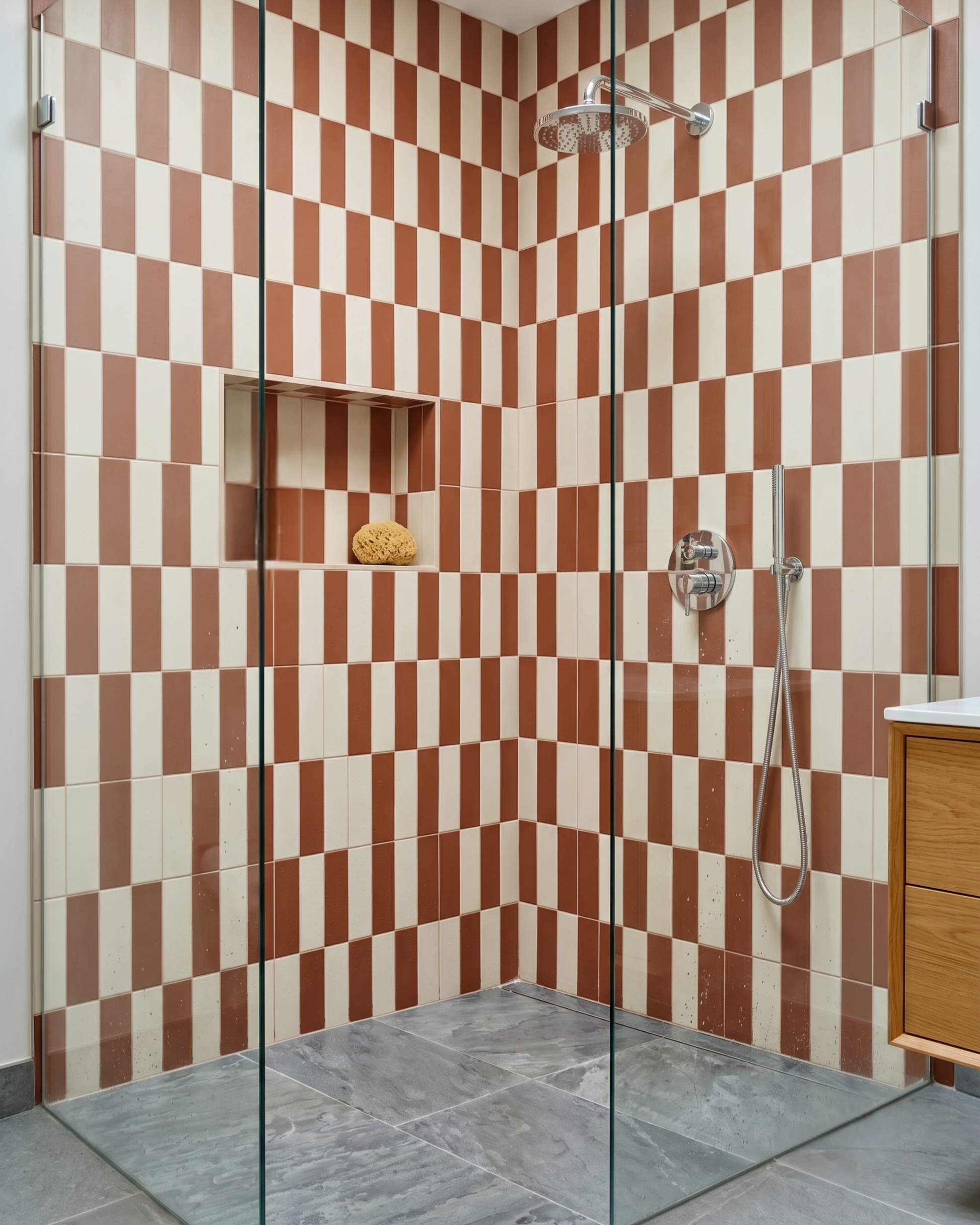 A modern walk-in shower stall with a brown and cream checkered tiled wall, a built-in niche with a sponge, gray stone floor tiles, a glass door, and a chrome shower head and handheld hose.