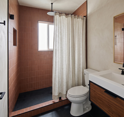 A modern bathroom interior featuring a walk-in shower with terracotta tiled walls and floor, a white textured shower curtain, a white toilet, a white sink with a wood cabinet, and a mirror on a beige wall.