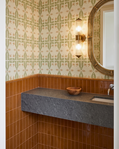A bathroom vanity corner with patterned wallpaper, warm brown tile wainscoting, a dark floating stone countertop, a wooden bowl, and a round mirror with a gold frame.