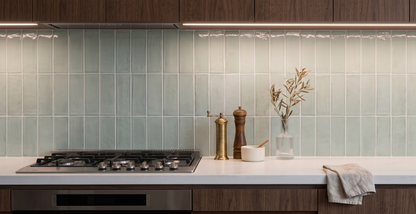A modern kitchen scene featuring a light sage green subway tile backsplash, dark wood cabinets, a white countertop with a built-in gas stove, and decorative items including a vase with dried branches, a salt and pepper grinder, and a folded cloth.