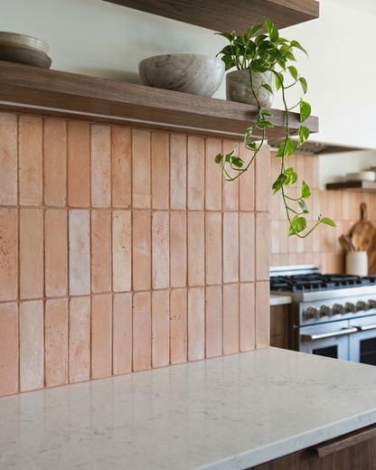 A kitchen backsplash made of vertical terracotta or peach-colored rectangular tiles, paired with a light stone countertop and dark wood floating shelves holding bowls and a trailing green plant.
