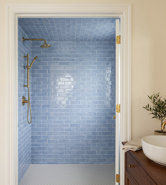 A bathroom interior showing a walk-in shower enclosure with glossy light blue subway tile walls and floor, brass shower hardware, a white door frame, and part of a wooden vanity with a white bowl sink on the right.