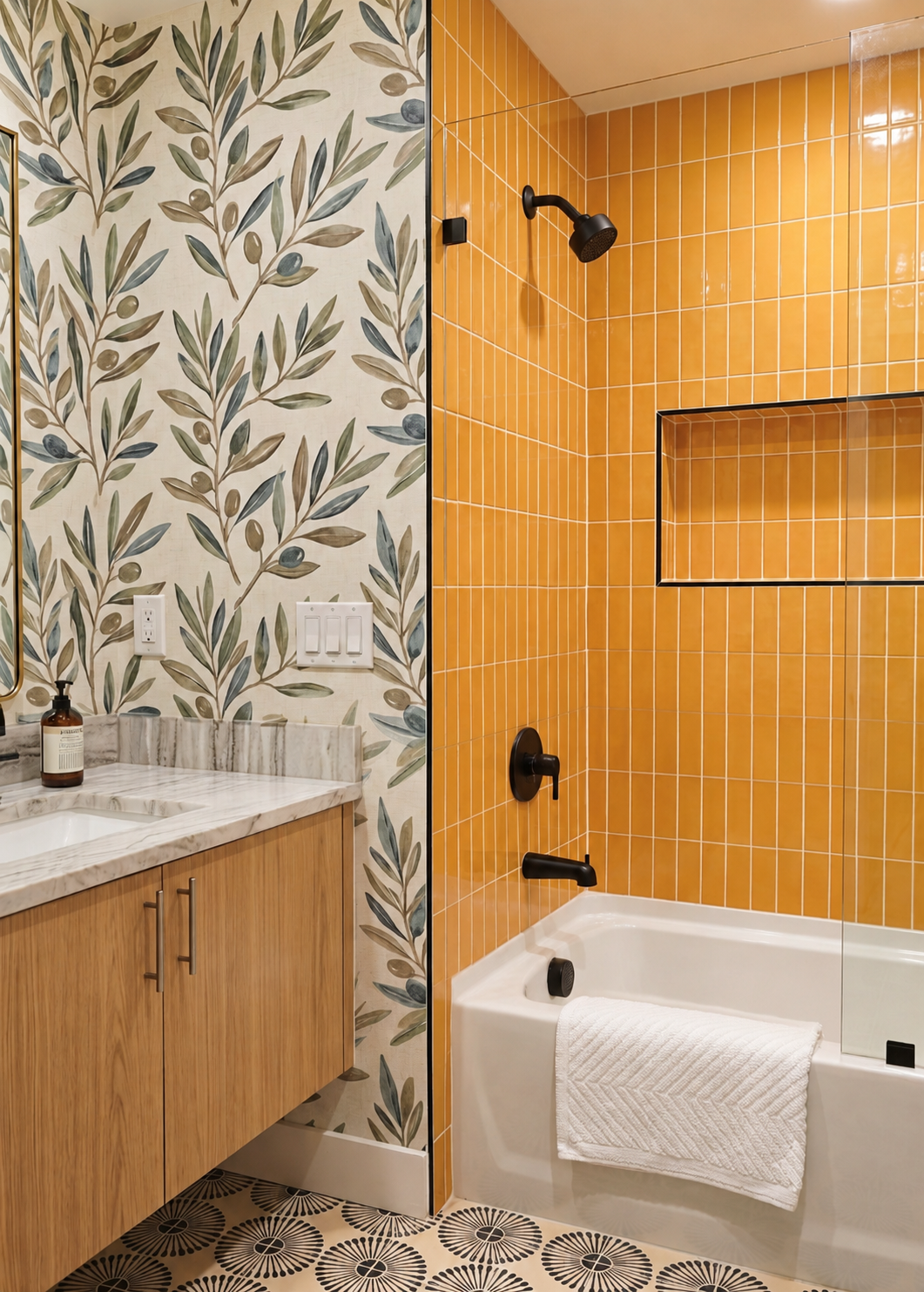 A modern bathroom interior featuring a white bathtub with a white towel, mustard yellow vertical tile wall, black fixtures, a marble countertop, and botanical pattern wallpaper.