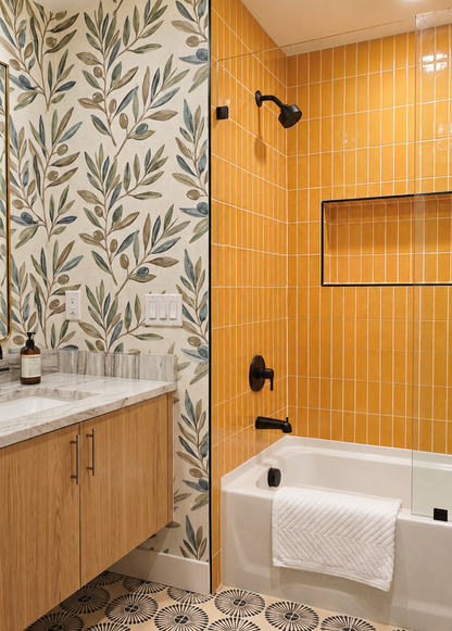 A modern bathroom interior featuring a white bathtub with a white towel, mustard yellow vertical tile wall, black fixtures, a marble countertop, and botanical pattern wallpaper.