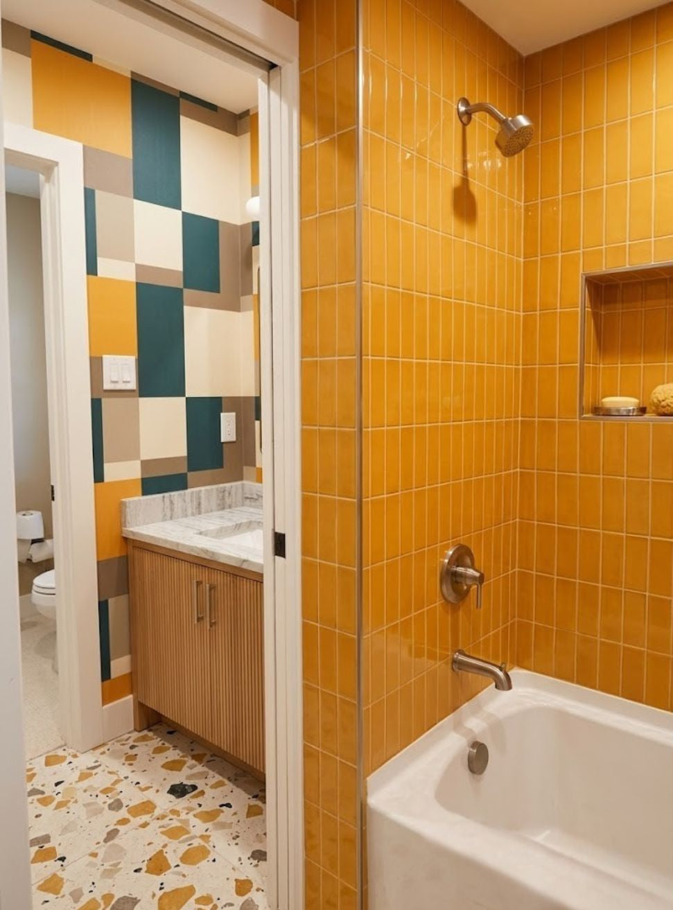 A bathroom interior with bright yellow-orange tiled walls, a white bathtub, a wall-mounted shower head, and a doorway opening to a vanity area with a geometric multicolor tile wall and terrazzo floor.