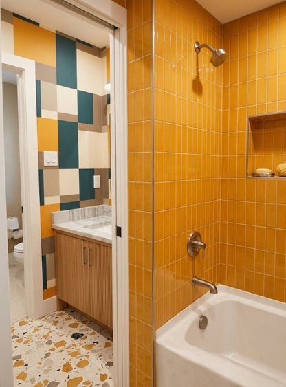A bathroom interior with bright yellow-orange tiled walls, a white bathtub, a wall-mounted shower head, and a doorway opening to a vanity area with a geometric multicolor tile wall and terrazzo floor.